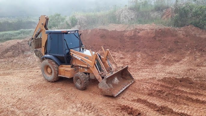 Retroescavadeira Case acertando o terreno de terra vermelha, com a lâmina baixada e o barranco cortado ao fundo.